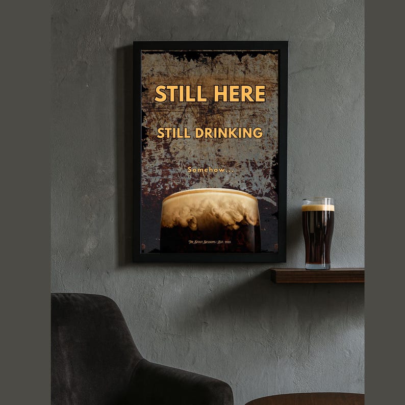 Close up framed Still Here Still Drinking wall art print in a moody interior setting, emphasizing distressed background textures, amber typography and stout beer imagery, suitable for pub rooms, kitchens, offices and modern rustic interior spaces