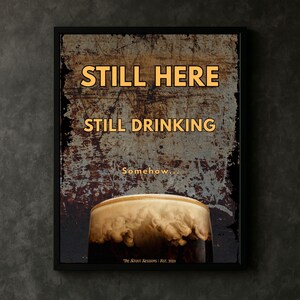 Close up framed Still Here Still Drinking wall art print in a moody interior setting, emphasizing distressed background textures, amber typography and stout beer imagery, suitable for pub rooms, kitchens, offices and modern rustic interior spaces