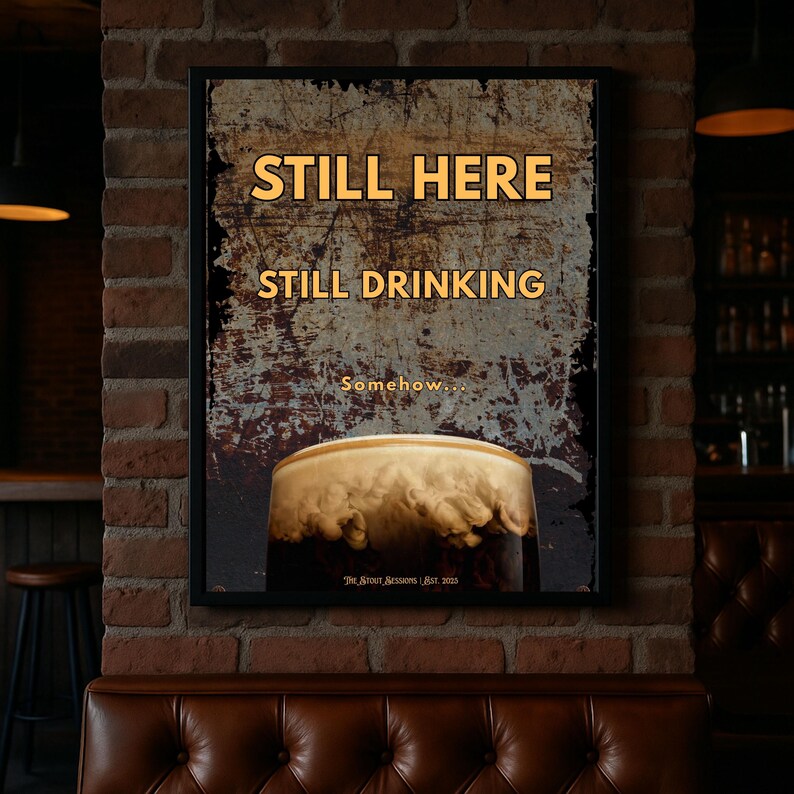 Framed Still Here Still Drinking wall art print shown in a dimly lit bar style interior with leather seating and warm tones, showcasing bold typography and stout inspired imagery, suitable for home bars, pubs, offices, studios and masculine interior decor