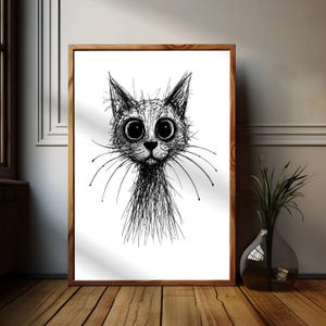 Könnte beinhalten: Ein gerahmter Schwarz-Weiß-Kunstdruck eines Katzenkopfes. Die Katze hat große, runde Augen und lange Schnurrhaare. Das Kunstwerk ist eine skizzenartige Zeichnung mit einem Holzrahmen. Der Druck wird auf einem Holzboden präsentiert.