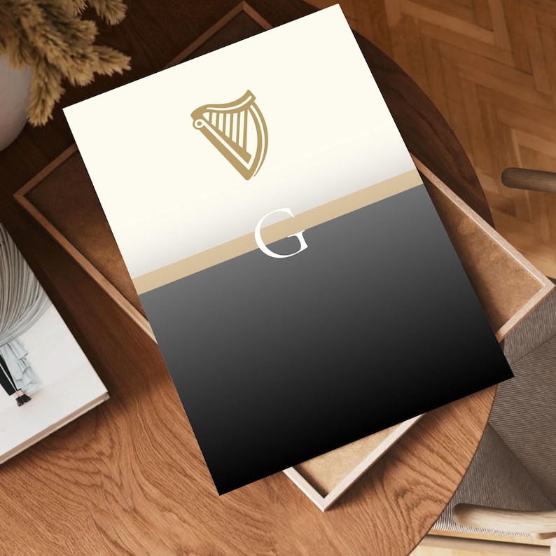 Guinness inspired “Split the G” poster art print laying flat on wooden desk surface, angled view showing framed minimalist black and cream pint design with golden harp logo, perfect Irish beer gift idea, stylish modern Guinness wall art, ideal for man caves, kitchens, pubs or living rooms, minimalist typography print, unique artwork for Irish décor, cultural and contemporary gift for Guinness lovers and beer enthusiasts.