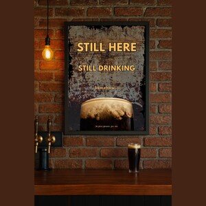 Still Here Still Drinking framed poster displayed against an exposed brick wall with warm ambient lighting, featuring distressed vintage textures and stout beer imagery, perfect for pub wall decor, bar interiors, man cave wall art and rustic or industrial home styling