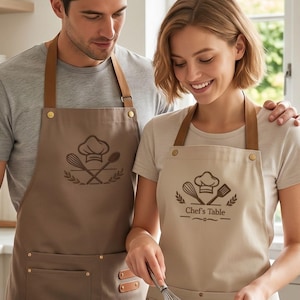 May include: Two aprons featuring brown leather-like straps and brass-colored hardware. The darker brown apron displays a chef hat and whisk graphic. The lighter beige apron features a chef hat, whisk, and spatula graphic with the text Chef's Table.