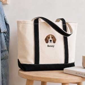 May include: A cream-coloured canvas tote bag with black trim and handles. The bag features an embroidered beagle dog face centred on the front, positioned above the name Benny stitched in black thread in a classic serif font.