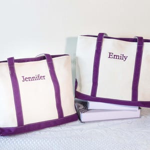 Op de afbeelding: Twee crèmekleurige canvas draagtassen met paarse accenten. Eén tas heeft de borduring "Jennifer", de andere "Emily". De tassen hebben paarse handvatten en randen.