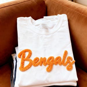 May include: A stack of folded white t-shirts with the word "Bengals" in orange, textured lettering. The shirts are stacked on top of other folded shirts in various colors, including navy and gray. The shirts are on a brown couch.