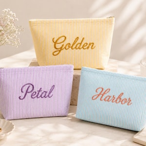 May include: Three zippered pouches made of seersucker fabric with white vertical stripes. The yellow pouch features the embroidered word Golden, the purple pouch features the word Petal, and the light blue pouch features the word Harbor in script.