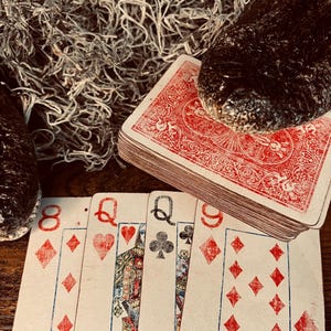 May include: A deck of playing cards with a red patterned back rests on a wooden surface, partially covered by a dark textured object. Four cards are fanned out in front: the eight of diamonds, queen of hearts, queen of clubs, and nine of diamonds.