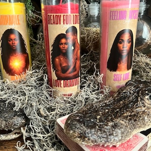 Puede incluir: Tres velas altas en frasco de vidrio con cera amarilla, roja y rosa. Cada vela tiene una etiqueta con un retrato y texto: Miracle, Ready For Love Love Drawing y Feeling Myself Self Love. Las velas están dispuestas con musgo seco.