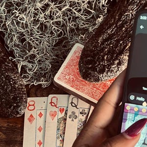 May include: A smartphone displaying a keyboard and the word Hello, positioned next to a deck of playing cards featuring an eight of diamonds and two queens, with two dark, textured, elongated objects resting on top of light-colored, wispy material.