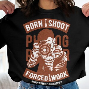 Op de afbeelding: Zwart sweatshirt met een bruine afbeelding van een persoon die een camera vasthoudt. De afbeelding zegt "Born to Shoot Photographer Forced to Work Independent Photographer".