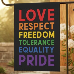 May include: A black garden flag displaying the words "LOVE, RESPECT, FREEDOM, TOLERANCE, EQUALITY, PRIDE" in rainbow colours. The flag is mounted on a black metal stand, set in a garden with white flowers and stone paving.