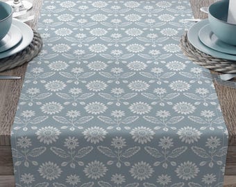 May include: A light blue table runner with a white floral pattern, set on a wooden table. The runner features a repeating design of stylized flowers and leaves. The table is set with plates, bowls, silverware, and wine glasses.