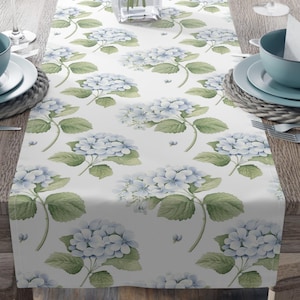 Könnte beinhalten: Ein weißer Tischläufer mit einem sich wiederholenden Muster aus blauen Hortensienblüten und grünen Blättern. Der Läufer liegt auf einem Holztisch, mit Gedecken und einer Vase mit rosa Lilien.