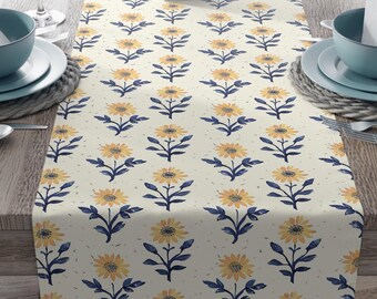 May include: Cream-colored table runner with a repeating pattern of yellow sunflowers and navy blue stems and leaves. The runner is set on a wooden table, with place settings and a vase of pink flowers.