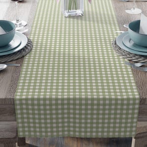 Puede incluir: Una mesa de comedor preparada para una comida, con un camino de mesa a cuadros verdes y blancos. La mesa está puesta con cuencos azules, platos blancos, cubiertos y copas de vino. Un jarrón de lirios rosas está en el centro.