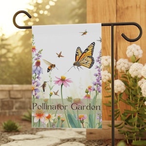 Puede incluir: Una bandera de jardín blanca con una mariposa colorida, abejas y colibríes volando alrededor de flores rosas y moradas. El texto "Pollinator Garden" está impreso en la bandera.