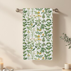 Puede incluir: Una toalla de mano blanca con un estampado botánico de hojas verdes y flores amarillas cuelga de una barra plateada. La toalla está en un ambiente de baño con una vela, un jarrón y piedras decorativas.