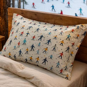 May include: A cream pillowcase featuring a repeating pattern of skiers in red, blue, and yellow. The pillow is placed on a bed with a wooden headboard. A snowy mountain scene with skiers is visible through a window, with a wooden cabin in the background.