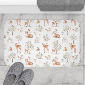 May include: A rectangular white bath mat with a repeating pattern of deer, trees, and mushrooms in shades of brown and beige. The mat is placed on a tiled floor, with a pair of gray slippers in the foreground.