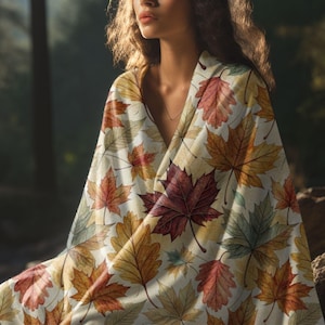 May include: A cream-coloured blanket featuring a pattern of autumn leaves in red, orange, yellow, and green. The blanket is draped over someone, creating a cosy and warm aesthetic. The leaves are detailed and realistic.