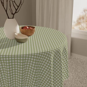 May include: A round tablecloth with a green and white checkered pattern covers a table. A white vase with decorative branches, a small bowl, and a basket of red apples are on the table. The tablecloth is made of a soft fabric.