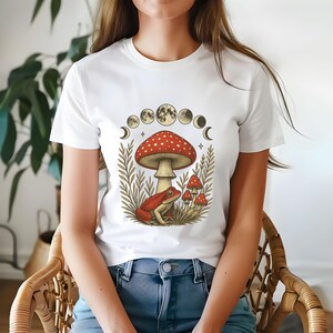 Könnte beinhalten: Weißes T-Shirt mit einem fantasievollen Design. Die Grafik zeigt einen roten Pilz, einen roten Frosch und kleinere Pilze, mit einer Reihe von Mondphasen und Blättern. Das T-Shirt hat einen Rundhalsausschnitt.