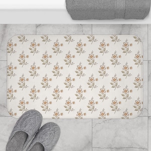 May include: A rectangular bath mat with a floral pattern in beige, brown, and orange. The mat features a repeating design of stylized flowers and leaves. Grey slippers are at the bottom of the mat. A grey towel is at the top.