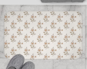May include: A rectangular bath mat with a floral pattern in shades of beige, brown, and orange. The mat features a repeating design of stylized flowers and leaves. Gray slippers are at the bottom of the mat. A gray towel is at the top.