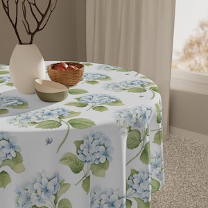 May include: A round white tablecloth with a pattern of blue hydrangea flowers and green leaves. The table is set with a white vase, a small woven basket with red apples, and two bowls. The tablecloth is draped over a round table.