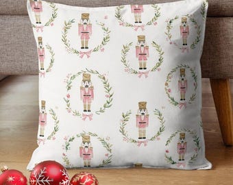 May include: White square pillow with a repeating pattern of pink nutcracker soldiers inside green wreaths with pink bows. The pillow is on a wooden surface with red and gold Christmas ornaments.