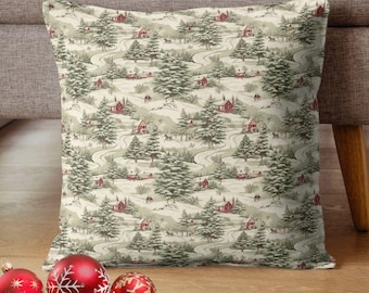 May include: Square decorative pillow with a winter landscape pattern featuring red houses, winding roads, and evergreen trees. The pillow is beige with a repeating pattern. Red and gold Christmas ornaments are in the foreground.
