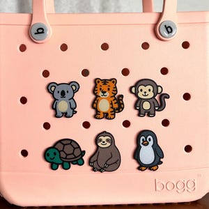 May include: A light pink tote bag with six animal-themed charms attached. The charms include a koala, tiger, monkey, turtle, sloth, and penguin. The bag has two handles and the brand name "bogg" is visible on the bottom.