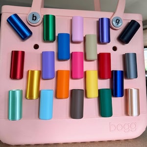 May include: A collection of colorful, cylindrical accessories in various shades, including blue, green, pink, and yellow. The items are arranged on a light pink tote bag, with the brand name "bogg" visible at the bottom.