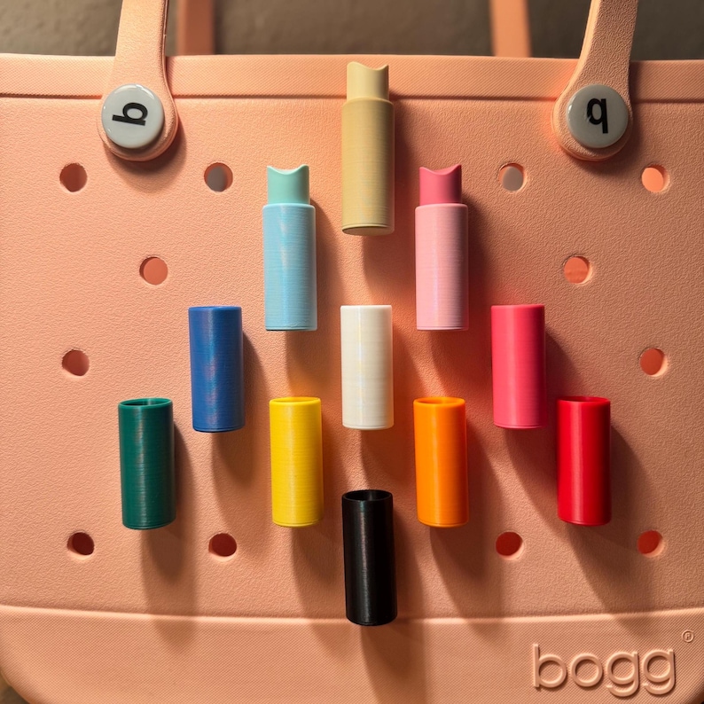 May include: A collection of colorful, cylindrical accessories, including green, blue, yellow, black, white, orange, pink, and red, are displayed against a peach-colored tote bag. The bag has a handle and the brand name "bogg" is visible.