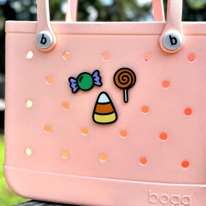May include: A light pink tote bag with a candy theme. The bag has a perforated design and is decorated with candy-themed charms, including a green wrapped candy, a lollipop, and a candy corn.