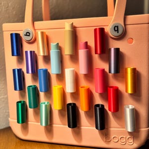 May include: A variety of colorful, cylindrical accessories are displayed on a light pink tote bag. The accessories come in various colors, including blue, green, yellow, and red. The bag has the word "bogg" printed on it.