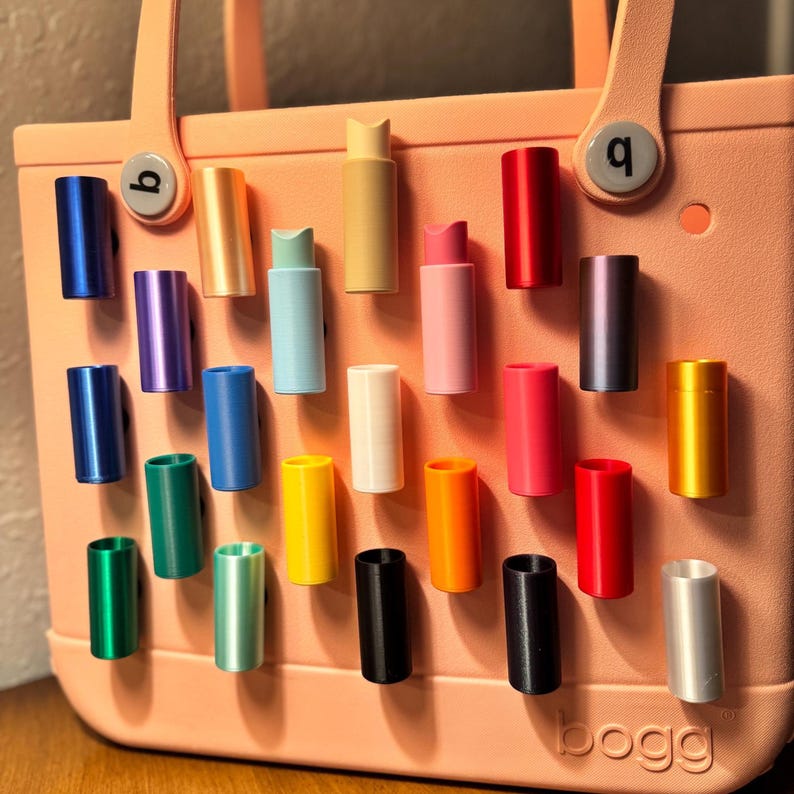 May include: A variety of colorful, cylindrical accessories are displayed on a light pink tote bag. The accessories come in various colors, including blue, green, yellow, and red. The bag has the word "bogg" printed on it.