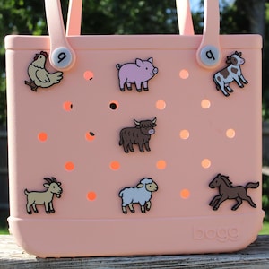 May include: A light pink tote bag with a variety of farm animal charms, including a chicken, pig, cow, bull, goat, sheep, and horse. The bag has a handle and the word "bogg" printed on the bottom.