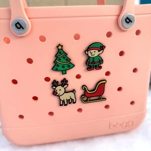 May include: A light pink tote bag with four Christmas-themed charms: a green Christmas tree, an elf, a reindeer, and a sleigh. The bag has a perforated design and a handle. The brand name "bogg" is visible on the bottom.