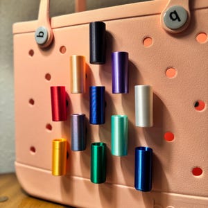 May include: A collection of colorful cylindrical accessories, including black, gold, red, blue, purple, and green, are displayed on a peach-colored tote bag. The accessories are arranged in a grid pattern, showcasing their various hues and metallic finishes.