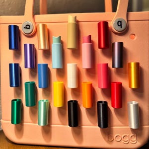 May include: A collection of colorful, cylindrical objects in various shades, including blue, green, yellow, and red, are displayed on a pink tote bag. The objects appear to be small containers or accessories, arranged in a grid-like pattern. The bag has the word "bogg" printed on it.