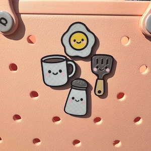 May include: A collection of cute, cartoon-style food-themed charms. The set includes a fried egg, coffee mug, salt shaker, and spatula, each with smiling faces. The charms are in black, white, yellow, and brown, and are set against a light pink background.