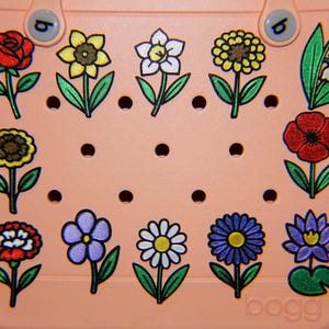May include: A peach-colored tote bag adorned with colorful floral designs. The flowers include a red rose, yellow daisy, white daffodil, and blue lotus. The bag has a perforated design and the word "bogg" is printed on the bottom.