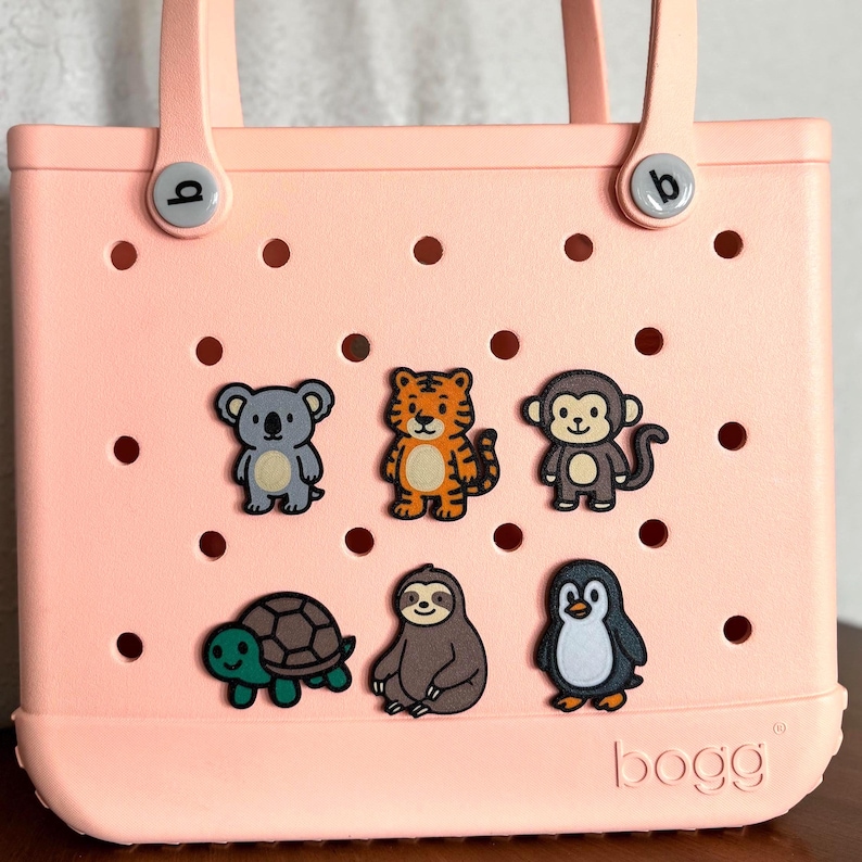 May include: A light pink tote bag with six animal-themed charms attached. The charms include a koala, tiger, monkey, turtle, sloth, and penguin. The bag has two handles and the brand name "bogg" is visible on the bottom.