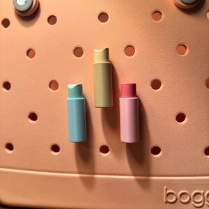 May include: Three colorful bag charms in pastel shades of blue, yellow, and pink. The cylindrical charms are displayed against a peach-colored background with circular cutouts. The word "bogg" is visible in the bottom right corner.