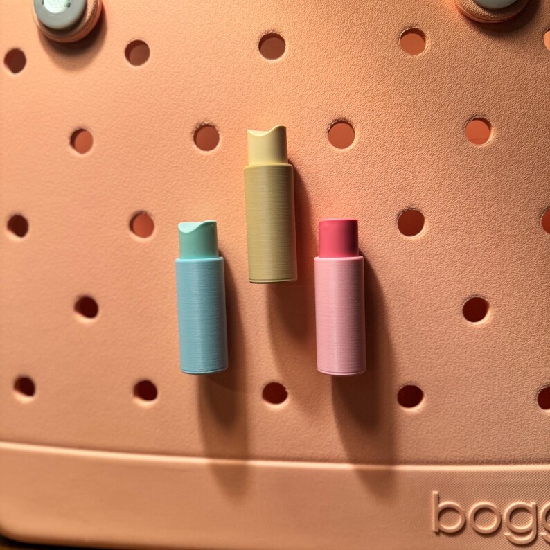 May include: Three colorful bag charms in pastel shades of blue, yellow, and pink. The cylindrical charms are displayed against a peach-colored background with circular cutouts. The word "bogg" is visible in the bottom right corner.