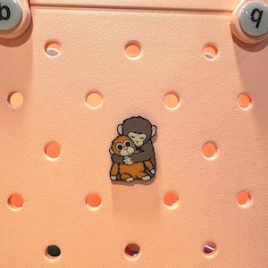 May include: A decorative charm featuring two hugging cartoon monkeys in shades of brown and orange. The charm is set against a light pink background with evenly spaced circular holes and two white circular buttons.