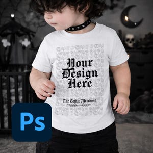 May include: White short-sleeved t-shirt with the text "Your Design Here" in a gothic font, over a floral pattern. The text "The Gothic Merchant" is below. The child is wearing a black choker and black trousers.