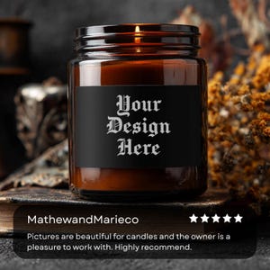 May include: A lit candle in a brown glass jar with a black label that reads "Your Design Here" in white gothic font. The jar sits on a dark surface. The background is blurred, with hints of dried flowers.